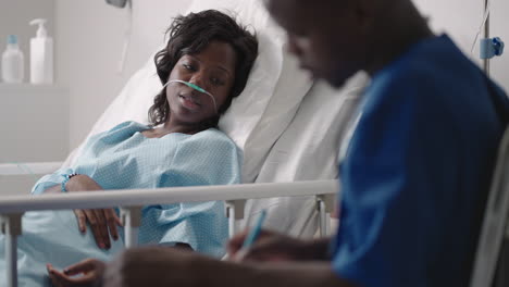 An-African-male-doctor-interviews-a-patient-lying-in-a-hospital-bed-with-an-oxygen-mask.-A-black-woman-lying-in-a-hospital-bed-describes-the-symptoms-to-the-doctor